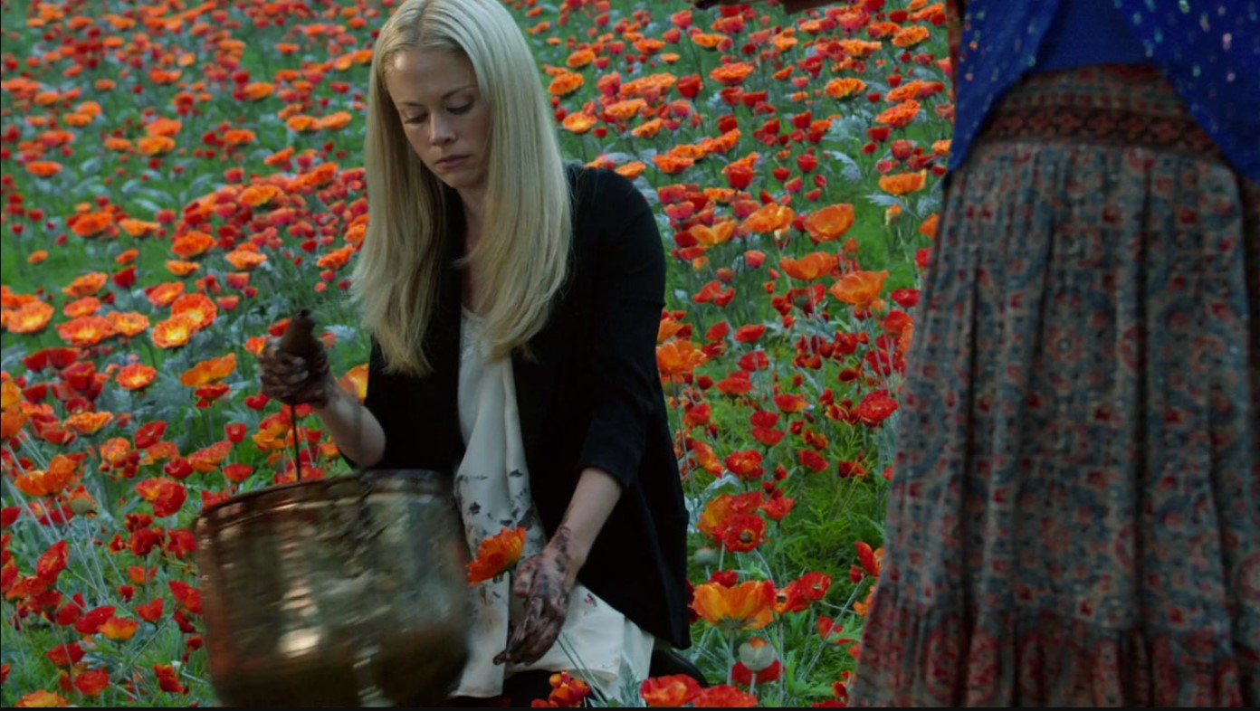 a blonde woman with bloodied hands sits on her knees and prepares to empty a metal bucket into a field of poppies